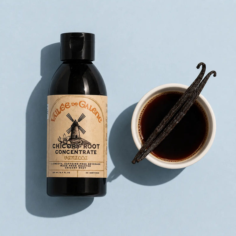 Bottle of chicory root concentrate with a cup of liquid and vanilla beans on a light blue background
